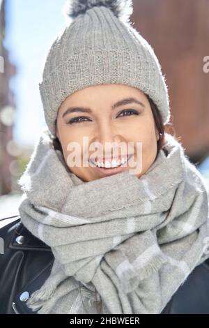 Primo piano di donna allegra in cappello di maglia grigio e sciarpa calda ed elegante sorridendo e guardando la macchina fotografica in una giornata di sole Foto Stock