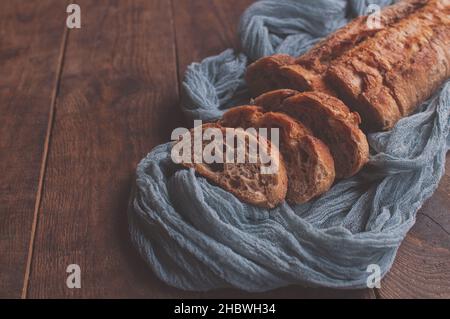 Pane fatto in casa cotto su un tavolo di legno con tovaglia di garza grigia, baguette fresche affettate su sfondo di legno Foto Stock