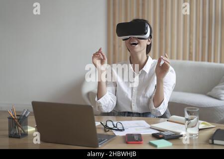 Lady imprenditore in visore VR seduto alla scrivania in ufficio a casa che interagisce con la visualizzazione 3D Foto Stock