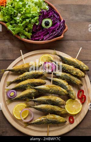 Sgombro di cavallo fritto, pesce istavrit bastoni di legno spiediti, su piatto di legno con insalata di stagione. Vista dall'alto di tavolo di legno. Foto Stock