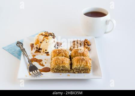 Tradizionale Baklava di noci turche in piatto quadrato con gelato alla vaniglia, noci e forchetta d'epoca. Servito con una tazza di tè su una superficie bianca Foto Stock