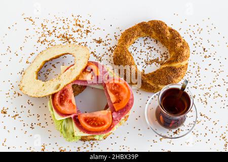 Tradizionale, croccante Bagel Turco, sandwich Simit con pomodoro, salame, foglia di lattuga e formaggio sulla superficie bianca con tè turco Foto Stock