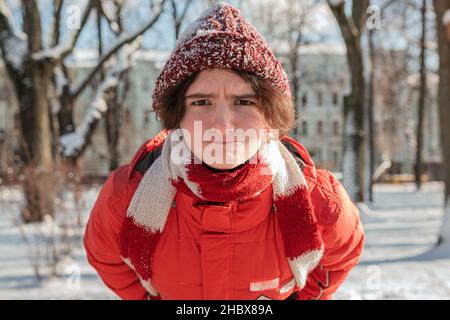 ragazza divertente adolescente in giacca calda giù e cappello e sciarpa in maglia con guance arrostbiten rosse e naso accigliato con sguardi espugnanti nella macchina fotografica Foto Stock