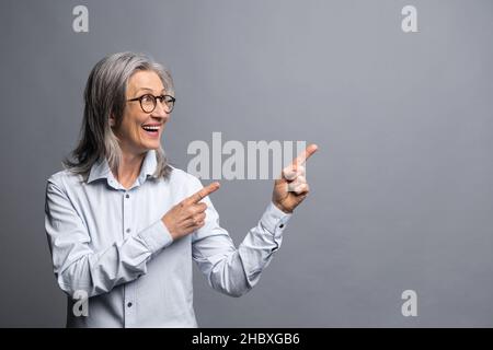 Fai pubblicità qui. Donna d'affari positiva indossando abiti di stile ufficiale puntando dito di distanza e prestando la vostra attenzione a spazio vuoto per la pubblicità. Studio interno girato isolato su sfondo grigio Foto Stock