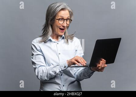 Sorprende la donna matura moderna usando il laptop isolato sul grigio. Donna anziana di tipizzazione imprenditore, donna di mezza età con capelli grigi in casual abbigliamento controllo e-mail Foto Stock