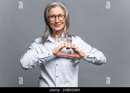 Ti amo. Ritratto di attraente donna matura romantica in piedi e facendo il cuore con le mani, mentre sorridendo giocosamente. Studio interno girato isolato su sfondo grigio Foto Stock