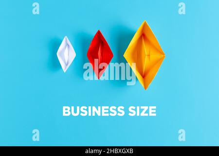 Concetto aziendale o aziendale. Tre barche di carta con dimensioni e colori diversi su sfondo blu con il testo business size. Foto Stock