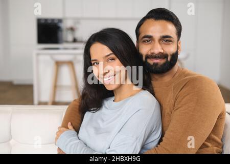Primo piano di felice coppia interrazziale in posa mentre si siede al divano con sfondo cucina. Felici i proprietari di un nuovo appartamento sorridente e abbracciato davanti alla fotocamera Foto Stock