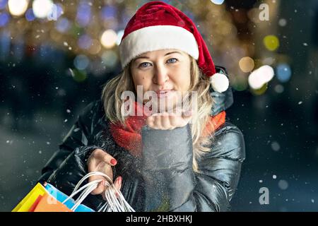 Shopping natalizio di una giovane donna. Sacchetti di carta colorati e luminosi con regali. Soffia i fiocchi di neve dalla mano. Foto Stock