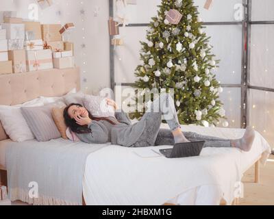 Giovane bella donna caucasica con capelli scuri in morbido maglione a maglia grigio usando il laptop sul letto in camera con albero di Natale. Decide le questioni relative alle prossime vacanze, ordini di consegna, effettua acquisti Foto Stock