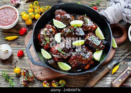 Vista dall'alto di una coscia di pollo piccante in una padella in ghisa su un tagliere di legno e sfondo rustico con ingredienti freschi. Foto Stock