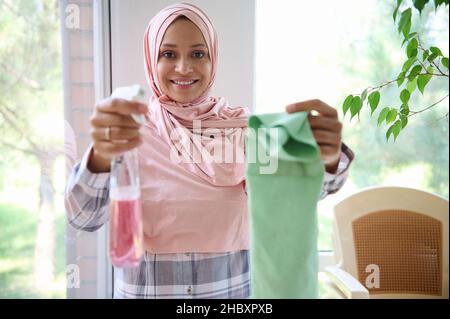 Affascinante musulmana araba bella donna in hijab sorride toothy sorriso guardando la macchina fotografica, tenendo un panno e detergente spray per il lavaggio delle finestre nella sua esterno Foto Stock