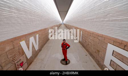 22 dicembre 2021, Sassonia, Chemnitz: L'artista Anke Neumann si trova nel nuovo tunnel pedonale della stazione centrale di Chemnitz. Lo stretto tunnel tra il centro della città e il quartiere Sonnenberg è stato migliorato, tra le altre cose, da un nuovo concetto di illuminazione. Inoltre, lettere bianche alte 1,40 metri sulle pareti formano una citazione dalla canzone 'Wie ich' della band Kraftklub: 'ICH wär'gern weniger wie ich, ein bisschen mehr so wie du'. Nella volgare, il sottopasso, precedentemente piuttosto scuro, lungo 220 metri, è comunemente conosciuto come il 'tubo germinale'. In due fasi di costruzione, il entra Foto Stock