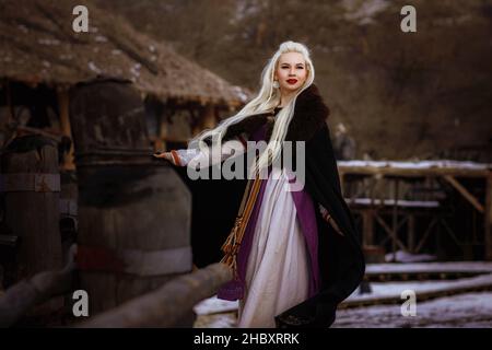 Bellissimo vichingo biondo vestito in un mantello nero sullo sfondo del castello. Bel set cinematografico - un antico villaggio vichingo Foto Stock