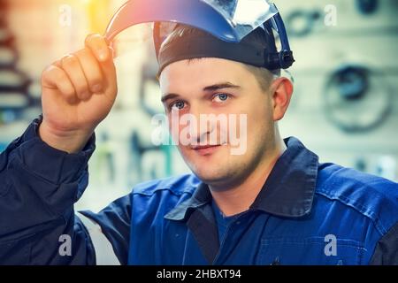 Ritratto di Metal Locksmith. Il giovane uomo caucasico guarda nella fotocamera da sotto visiera di maschera protettiva. Vero lavoratore in officina. Foto Stock