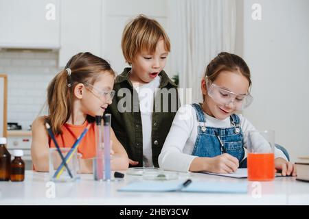 I capretti piccoli felici che fanno il progetto domestico di scienza, la ragazza sta documentando il processo. Ragazze che indossano occhiali protettivi, ragazzo facendo volto sorpreso. Prodotti chimici Foto Stock