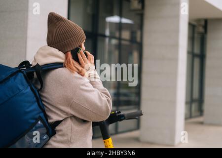 Vista posteriore del corriere femminile irriconoscibile che parla al telefono con il ristorante o con il cliente, consegnando l'ordine su scooter elettrico. Foto Stock