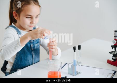 La bambina paziente che fa il progetto di scienza domestica, mescolando i liquidi rossi e blu in una fiasca. Ha vetreria chimica e microscopio Foto Stock
