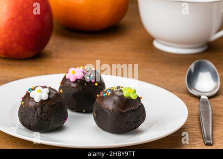 Palline di cioccolato o palline di cioccolato condite con spruzzi di arcobaleno multicolore e fiori di zucchero colorati in un piatto di ceramica bianca su un tavolo di legno con appl rosso Foto Stock