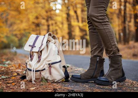 Donna Hitchhiker con zaino in attesa di una macchina sulla strada nella foresta in autunno. Donna che indossa scarpe scamosciate. Foto Stock