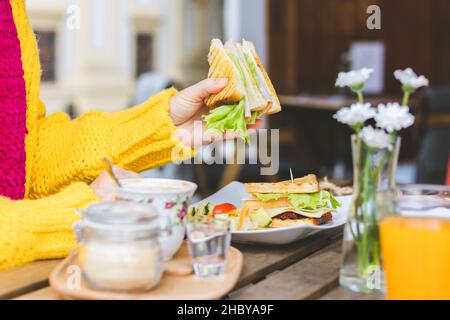 Donna che fa colazione in un caffè all'aperto in via città. Sandwich e caffè serviti al tavolo. Donna che indossa maglione di lana godendosi la mattina al ristorante Foto Stock
