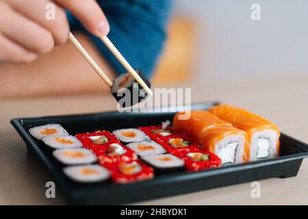 Vista frontale ravvicinata mani di un giovane irriconoscibile che tiene sushi con bacchette seduti alla scrivania. Foto Stock