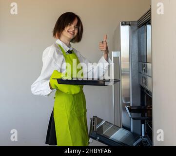 donna in piedi in grembiule verde tenendo una teglia da forno in una mano e facendo un segno giusto con l'altra. Sfondo semplice Foto Stock