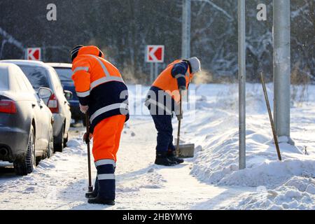 Rimozione della neve in città d'inverno alle basse temperature. Due uomini lavoratori in uniforme con una pala e crowbar pulizia della strada vicino al parcheggio auto Foto Stock