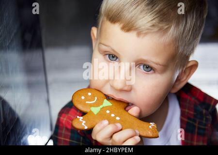 Il capretto caucasico carino mangia il pan di zenzero e fa un'espressione divertente del volto. Cibo di Natale. Casa di Natale suggestiva ed estetica per la vigilia di Capodanno. Ragazzo allegro. Gioioso, di buon umore. Foto Stock