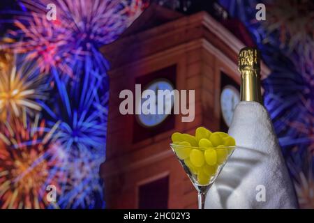Bicchiere con uva sullo sfondo di una bottiglia di champagne sfocata, torre dell'orologio alle dodici e fuochi d'artificio. Tradizione spagnola di mangiare dodici g. Foto Stock