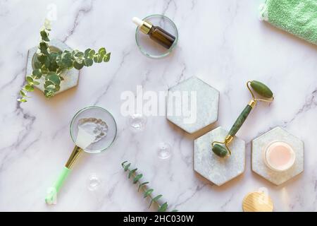 Moisturizer fatto a mano e rullo per il viso in giada verde. Asciugamano di cotone. Foglie di eucalipto esotico e gocce d'acqua su sfondo di marmo bianco con pietra Foto Stock