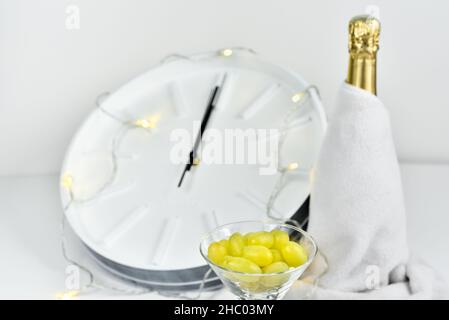 Uva in un bicchiere sullo sfondo di un orologio sfocato e una bottiglia di champagne. La tradizione spagnola di mangiare dodici uve per festeggiare l'anno nuovo. Foto Stock