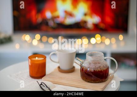 Foto di un luogo accogliente con tazza di tè, teiera, candela e libro con bicchieri con imitazione camino in tv sullo sfondo Foto Stock
