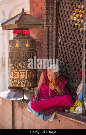 Una anziana suora buddista siede su una grande ruota di preghiera in stile tibetano presso il Boudhanath Stupa, Kathmandu, Nepal. Foto Stock