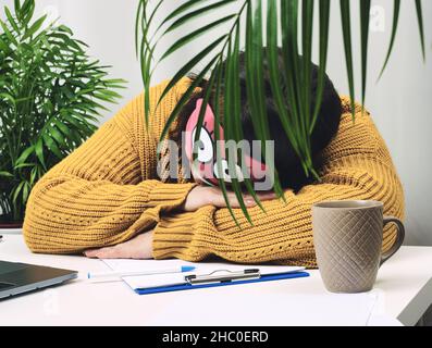 donna di 40 anni in un maglione arancione siede ad un tavolo con una maschera di sonno sul suo viso. La testa è sul tavolo, stanca e annoiato. Burnout e lavoro eccessivo Foto Stock