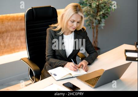 Successo Smart influente maturo concentrato fiducioso caucasico bionda donna d'affari, broker o esperto finanziario, seduto a tavola in ufficio con notebook, prendendo appunti, sviluppa la strategia di business Foto Stock