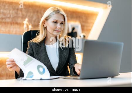 Caucasica maturo influente e di successo smart business donna, analista finanziario, manager, seduto a un laptop, studiare documenti, analizzare diagrammi e pianificare la strategia finanziaria Foto Stock