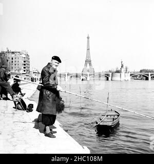 Parigi pescatori sulla Senna guardando la macchina fotografica - Pont Mirabeau, marzo 1945. Il suo vestito tradizionale include un cappotto di pelle, berretto e pantaloni a forma di culotte sopra la sox di lana. Un cannello da pesca è appeso alla spalla. La sua canna da pesca è molto più lunga di quanto sia alto. Pali vicino sul fiume sono barche a remi e altri pescatori. La Statua della libertà Petite è di fronte alla macchina fotografica da Pont de Grenelle. Alle spalle si vedono il Pont de Bir Hakeim e la Torre Eiffel. Foto Stock