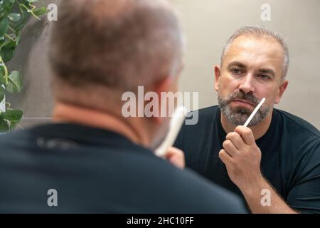 Un bell'uomo di mezza età che gli spazzolava la barba la mattina in bagno guardando nello specchio Foto Stock