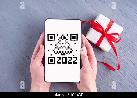 Mani che tengono uno smartphone, un tablet, un telefono cellulare con uno schermo qr in forma di albero di Natale data 2022 e una confezione regalo su sfondo grigio. Foto Stock