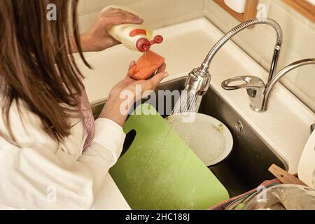 La donna versa il detersivo sulla spugna per lavare i piatti sporchi con acqua corrente nel moderno lavandino in cucina luce vista estrema ravvicinata Foto Stock