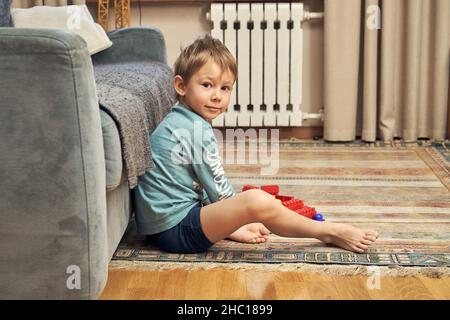 Curioso ragazzo preschooler gioca con dettagli costruttore in plastica colorata seduta su tappeto vicino divano in soggiorno luminoso a casa Foto Stock