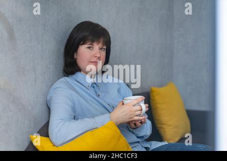 Bella giovane donna con tazza di caffè è seduta sul divano a casa. Donna che riposa durante la pausa pranzo. Rilassatevi con il tè. Foto Stock