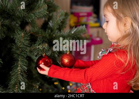 Piccola bella ragazza bionda in rosso in piedi vicino all'albero di Natale e toccando due bolle decorative rosse sull'albero che li guarda. Foto Stock