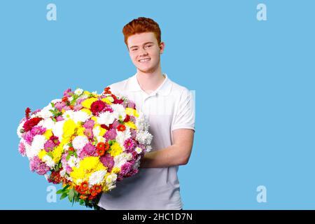 Giovane uomo capelli rossi felice tenendo un grande bouquet di fiori nelle sue mani su sfondo blu, copia spazio. Foto Stock