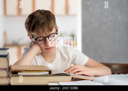 Ragazzo stanco in occhiali legge libri. Il bambino sta facendo i compiti molto. Homeschooling. Apprendimento a distanza Foto Stock