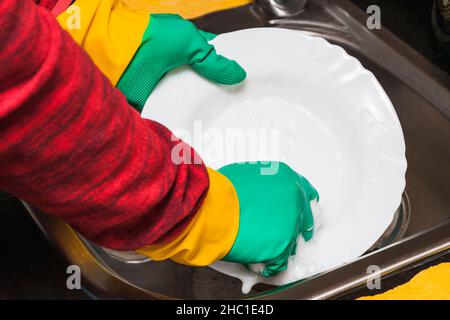 Primo piano delle mani di una persona che indossa guanti di gomma verdi e gialli strofinando un piatto di porcellana bianca con un tampone blu in un lavandino usando soa Foto Stock