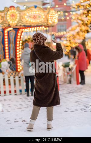 Donna in pelliccia cappotto, cappello panama al mercato di Natale di Tallinn fotografando con il cellulare Foto Stock