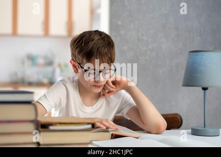 Ragazzo in occhiali legge libri mentre si siede in casa. Il bambino sta facendo i compiti. Homeschooling. Apprendimento a distanza Foto Stock