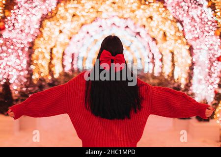 Giovane donna bruna in un maglione intimo lavorato a maglia e un arco rosso nei suoi capelli sullo sfondo di Natale luccicante luci rosa dorato. Vista posteriore Foto Stock